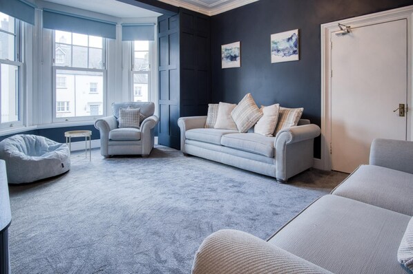 A living room with a grey sofa, a grey armchair, and a bean bag chair. There are two windows with blue shutters, a door, and two framed pictures on the wall. - The Lanterns - 9 Bedroom - Sleeps 17 - Saundersfoot Village Centre (Saundersfoot)