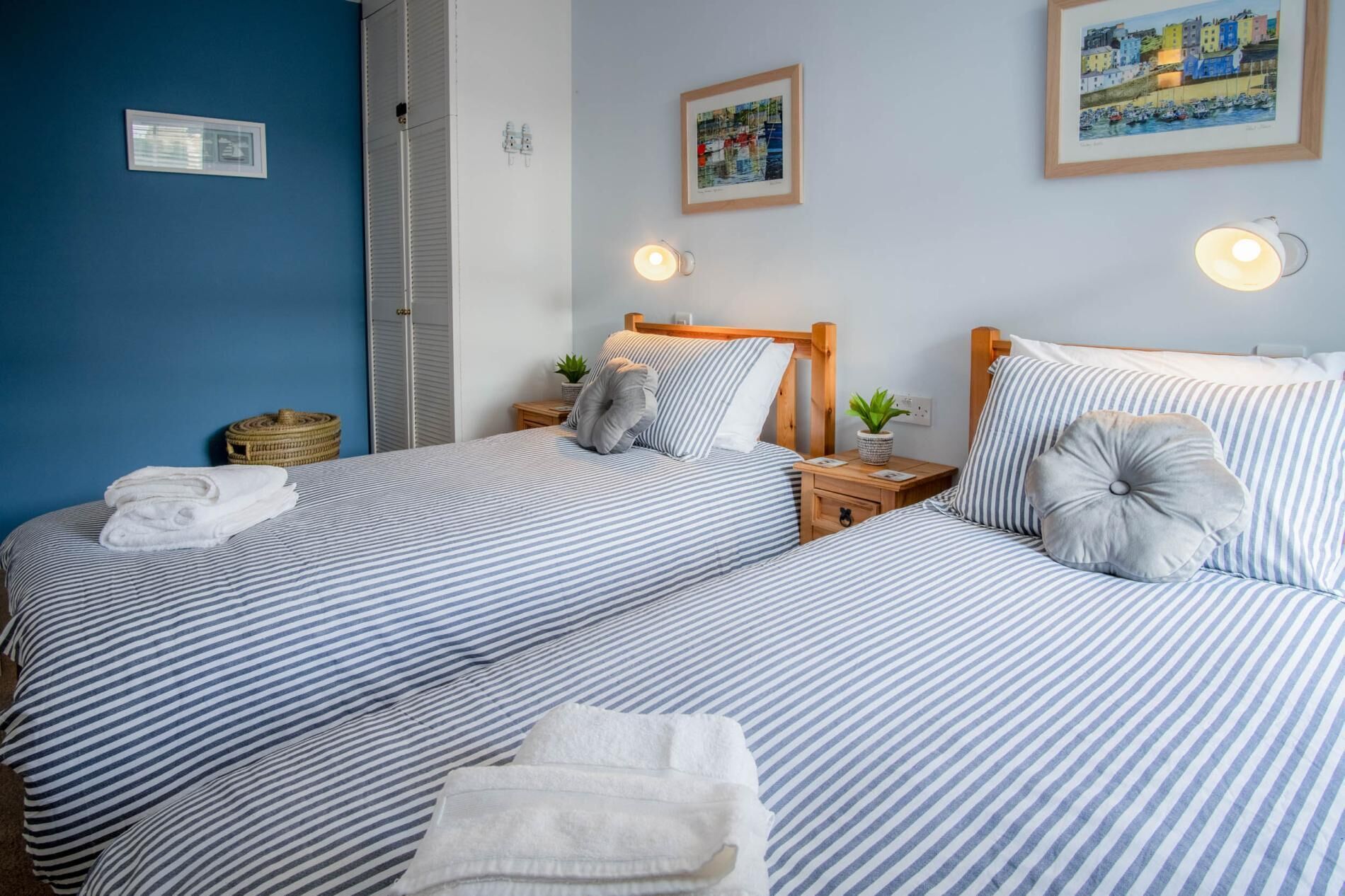 A room with two beds, each with striped bedding and pillows, a wooden headboard, bedside tables with lamps, and a framed painting on the wall.