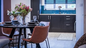 Un comedor con una mesa dispuesta para cuatro personas, un jarrón de flores y vista a la cocina a través de una ventana.