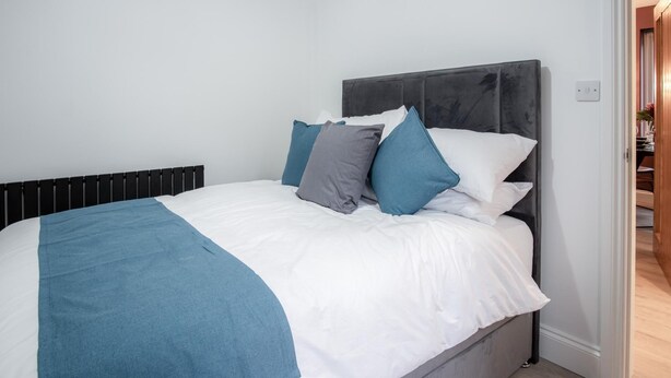 Una cama bien hecha con un cabecero gris, ropa de cama blanca y azul, y varios almohadones en tonos azules y grises.
