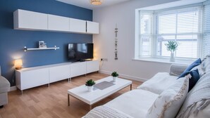 A modern living room with a TV, white furniture, a coffee table, and a window with blinds.