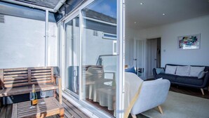 Een moderne woonkamer met een grote schuifglazen deur die leidt naar een balkon, een bank, een klein tafeltje met een fles en glas, en een bed met een decoratief beddegoed.
