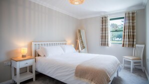 A bedroom with a bed, bedside table, lamp, chair, window with curtains, and a mirror. - 3 St Bride s Bay View - 3 Bedroom - Broad Haven (Haverfordwest)