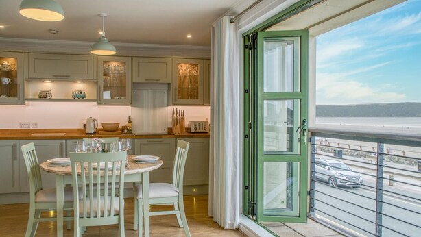 A kitchen with wooden cabinets, a dining table with chairs, and a view of a beach through a large window. - 3 St Bride s Bay View - 3 Bedroom - Broad Haven (Haverfordwest)