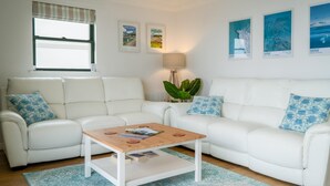 A modern living room with a white sofa set, a wooden coffee table, and a lamp. There are framed pictures on the wall and a window with blinds. - 3 St Bride s Bay View - 3 Bedroom - Broad Haven (Haverfordwest)