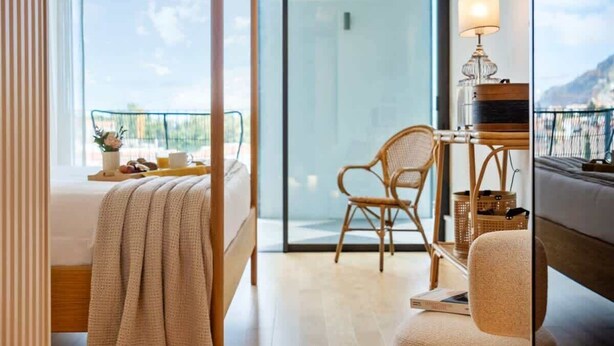 A hotel room with a bed, a chair, a table with a lamp, and a view of the outdoors. - Luxury 5-Star Stay at Savoy Insular (Funchal)