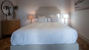 Une chambre à coucher bien rangée, avec un grand lit, deux tables de chevet avec des lampes, une petite table en bois et un miroir au mur.