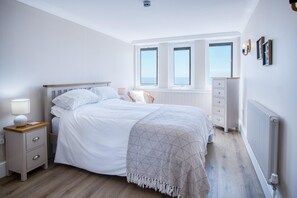 Une chambre à coucher comprenant un lit, une table de chevet, une commode et une vue sur la mer par les fenêtres.