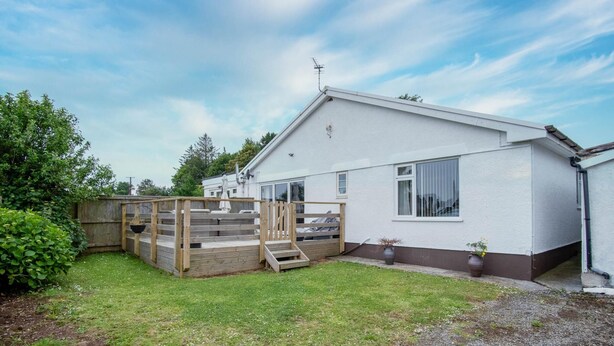 A single-story house with a white exterior, a wooden deck, and a small garden area. - Ridgeway Meadow Bungalow - 3 Bedroom - Manorbier (Tenby)