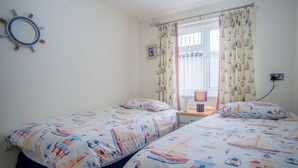 A bedroom with two beds, nautical-themed bedding, a window with curtains, a bedside lamp, and a wall-mounted mirror. - Ridgeway Meadow Bungalow - 3 Bedroom - Manorbier (Tenby)