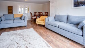 A living room with two grey sofas, a yellow armchair, a wooden dining table with chairs, and a white rug. - Ridgeway Meadow Bungalow - 3 Bedroom - Manorbier (Tenby)