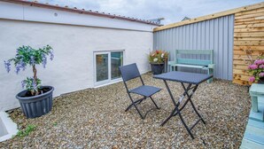 A small patio area with a white wall, a wooden bench, a table, and a potted plant. - Clouds - 1 Bedroom Holiday Home - Milford Haven (Milford Haven)