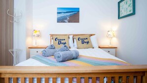 Cama bem arrumada com duas almofadas com a inscrição "Ocean Days", vista para a praia e um quadro na parede.