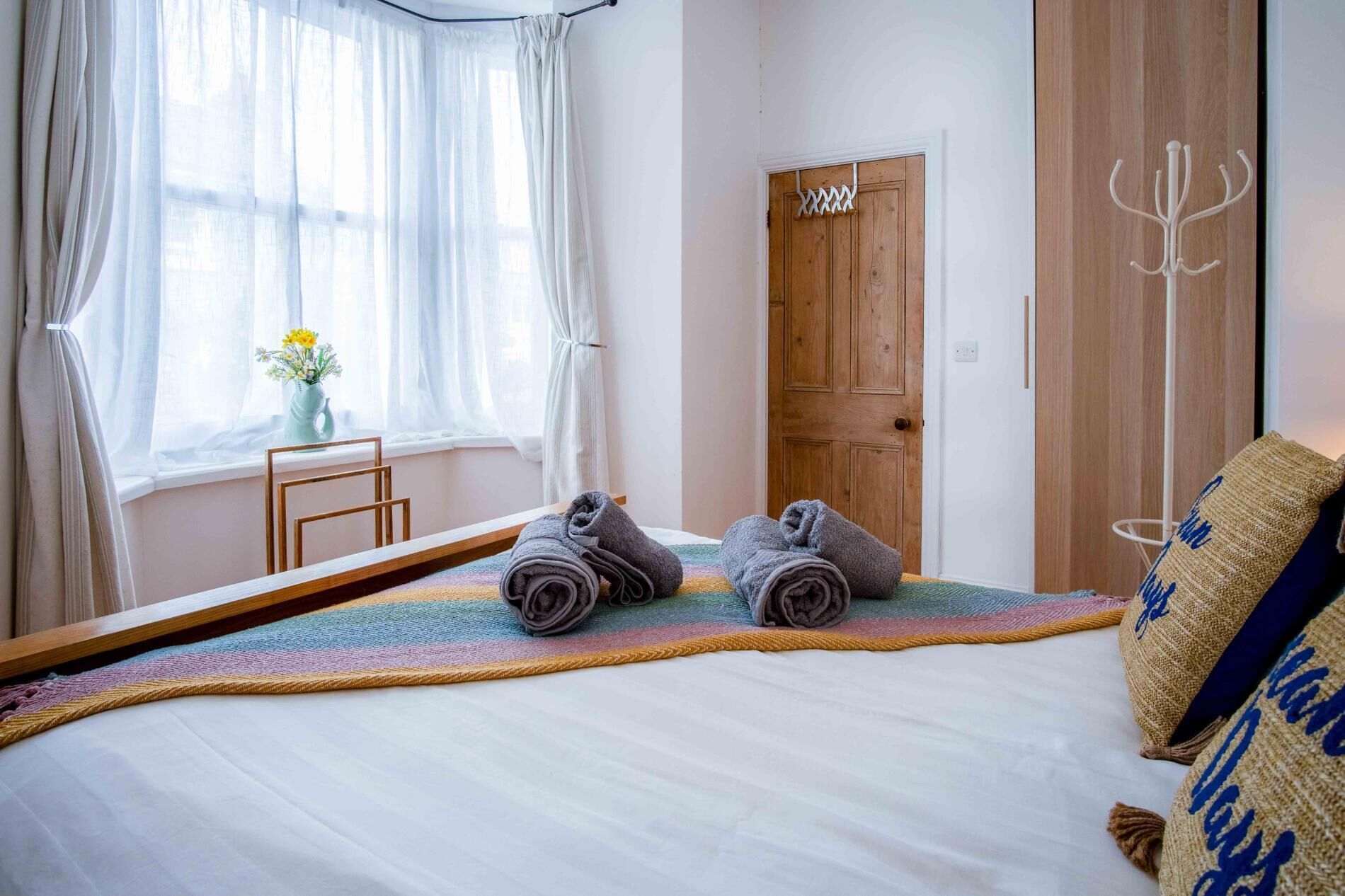 A neatly made bed with rolled towels, a wooden door, and a window with sheer curtains.