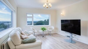 A modern living room with a sofa, two chairs, a TV, and a view of the city through large windows. - Longview - 4 Bedroom Holiday Home - Pembroke Dock (Pembroke Dock)
