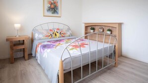 A bedroom with a metal-framed bed, a bedside table with a lamp, a wooden nightstand, and a wall-mounted shelf with two small potted plants. - Longview - 4 Bedroom Holiday Home - Pembroke Dock (Pembroke Dock)
