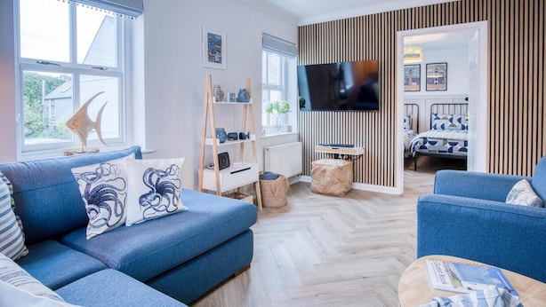 Ein modernes Wohnzimmer mit einer blauen Sofagarnitur, einem Flachbildfernseher, der an einer Holzwand angebracht ist, und einem Bett mit einem geometrisch gemusterten Überwurf im Hintergrund.