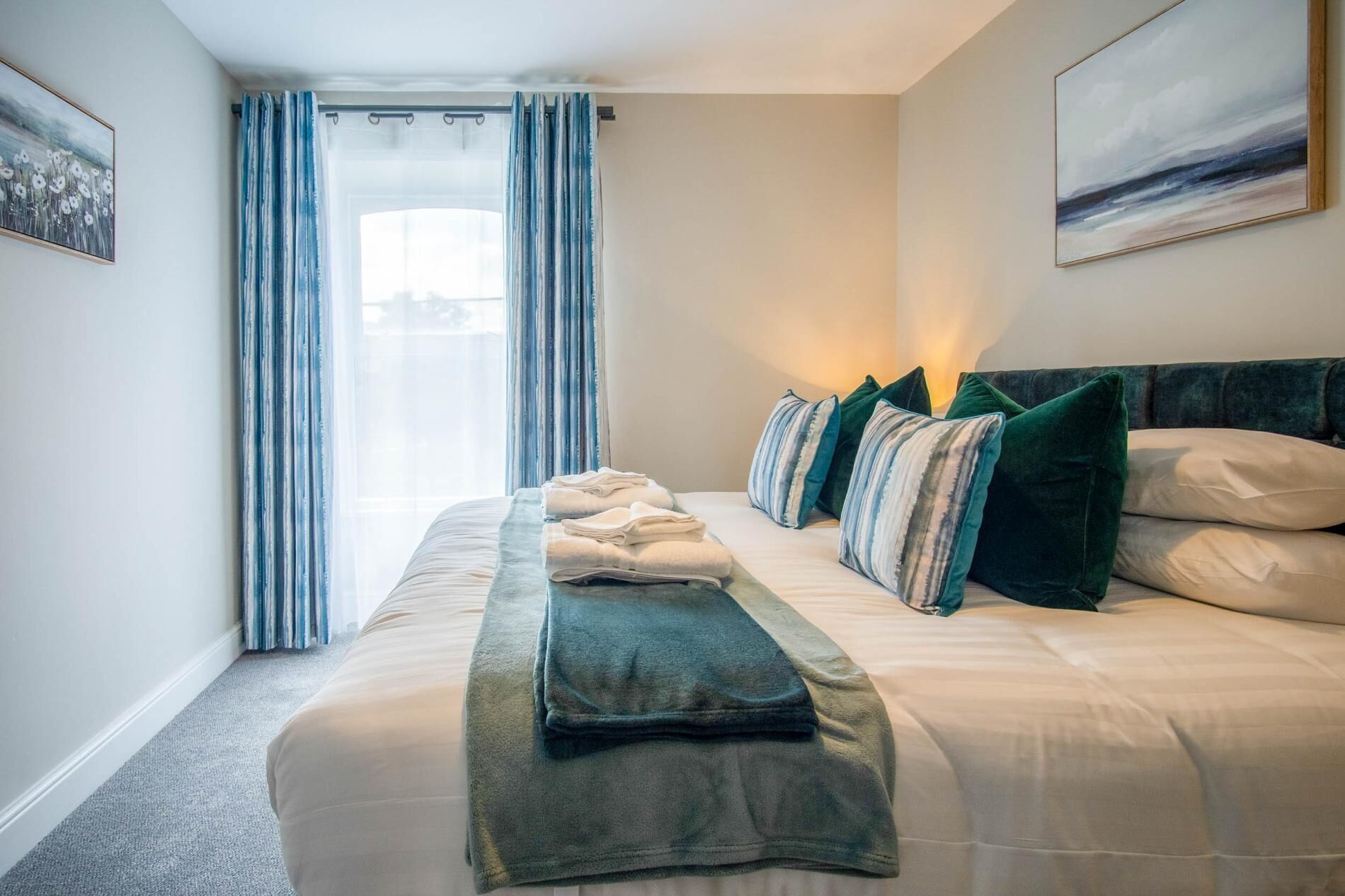 A neatly made bed with pillows and a blanket, a window with curtains, and a painting on the wall.