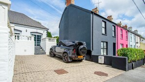 Un coche descapotable negro estacionado en una entrada, con una casa blanca y casas urbanas coloridas al fondo.
