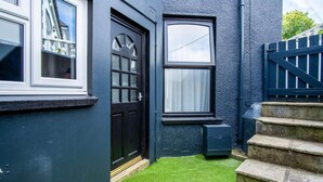 Una puerta negra con un ventanal encima, una ventana pequeña con cortina, y una escalera de piedra que sube a una reja.
