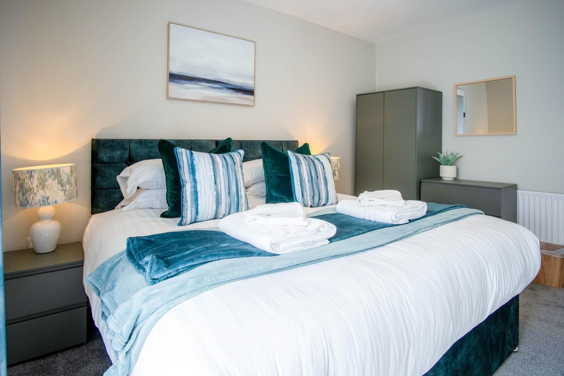 A neatly made bed with white linens, a teal throw blanket, and matching pillows. There is a bedside lamp, a small potted plant, and a mirror on the nightstand.