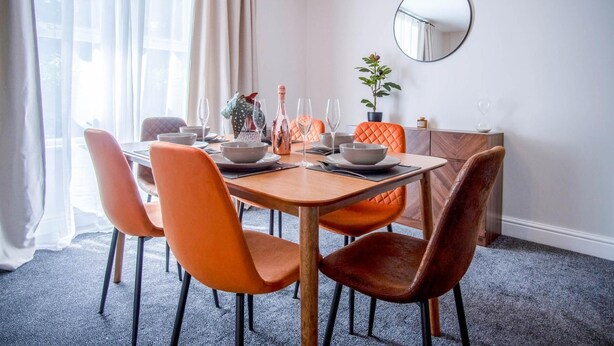 Un comedor con mesa de madera dispuesta para cuatro personas, rodeada de sillas anaranjadas. Un espejo cuelga de la pared y hay una planta sobre un mueble.