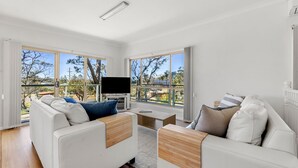 A spacious living room with a large TV, comfortable seating, and large windows offering a view of the outdoors. - Ocean Echoes - Pet Friendly with Ocean Glimpses (Culburra Beach)