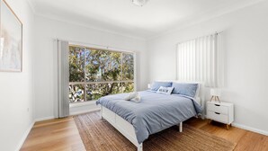 A bedroom with a bed, a nightstand, a window with blinds, and a view of trees outside. - Ocean Echoes - Pet Friendly with Ocean Glimpses (Culburra Beach)