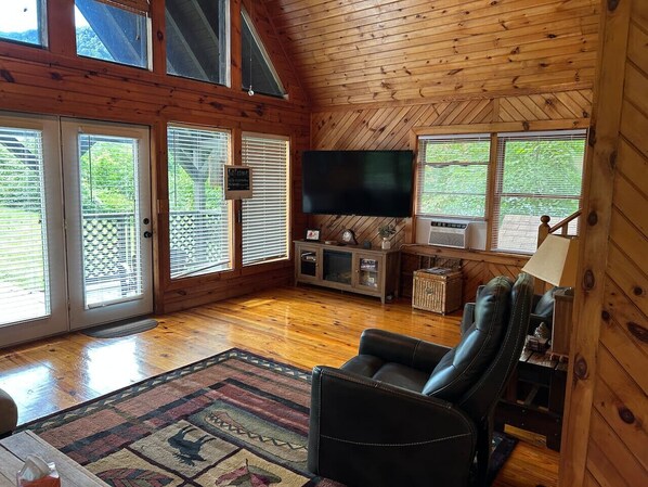 A cozy cabin interior with a wooden ceiling, a flat-screen TV, a leather chair, and a large window with blinds.