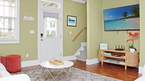 A living room with a white door, a staircase, a bookshelf, and a painting of a beach scene. - Seabbatical | Pet-Friendly Cottage | Screened Decks + Community Pools 🐾 (Port St. Joe)