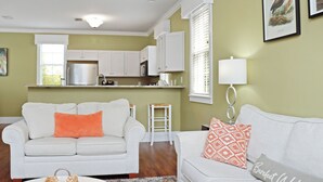 A living room with a white sofa, a coffee table, and a kitchen area with white cabinets and a refrigerator. - Seabbatical | Pet-Friendly Cottage | Screened Decks + Community Pools 🐾 (Port St. Joe)
