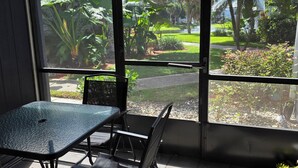 Una habitación con una mesa y sillas de vidrio, ventanas amplias con vistas a un jardín y árboles.