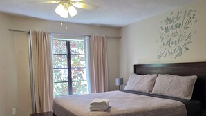 Un dormitorio con cama, ventilador de techo, ventana con cortinas y una pared decorada con un adhesivo de temática natural.
