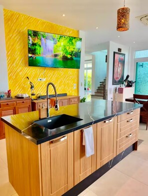 Une cuisine moderne dotée d’un grand îlot central, d’un téléviseur fixé au mur et d’un luminaire suspendu.