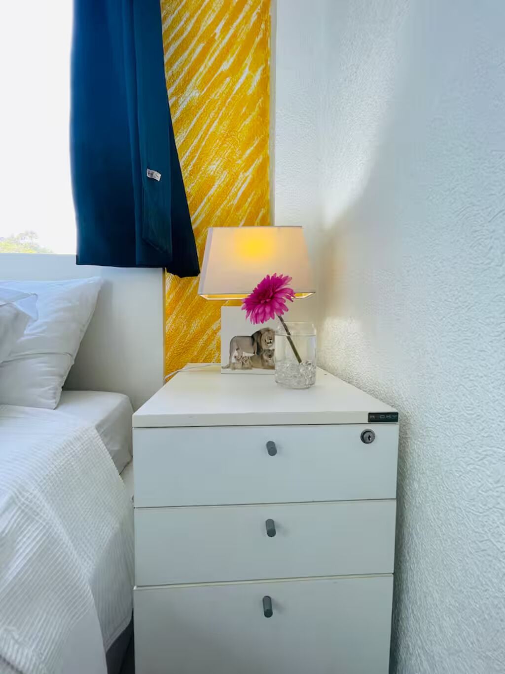 En säng med vita sängkläder, ett vitt nattduksbord med lådor, en lampa och en dekorativ prydnad med en rosa blomma.