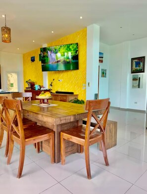A dining area with a wooden table and chairs, a TV mounted on a yellow wall, and a tiled floor. - Sea View Luxury Mansion • Pool & Jacuzzi (Koh Chang)