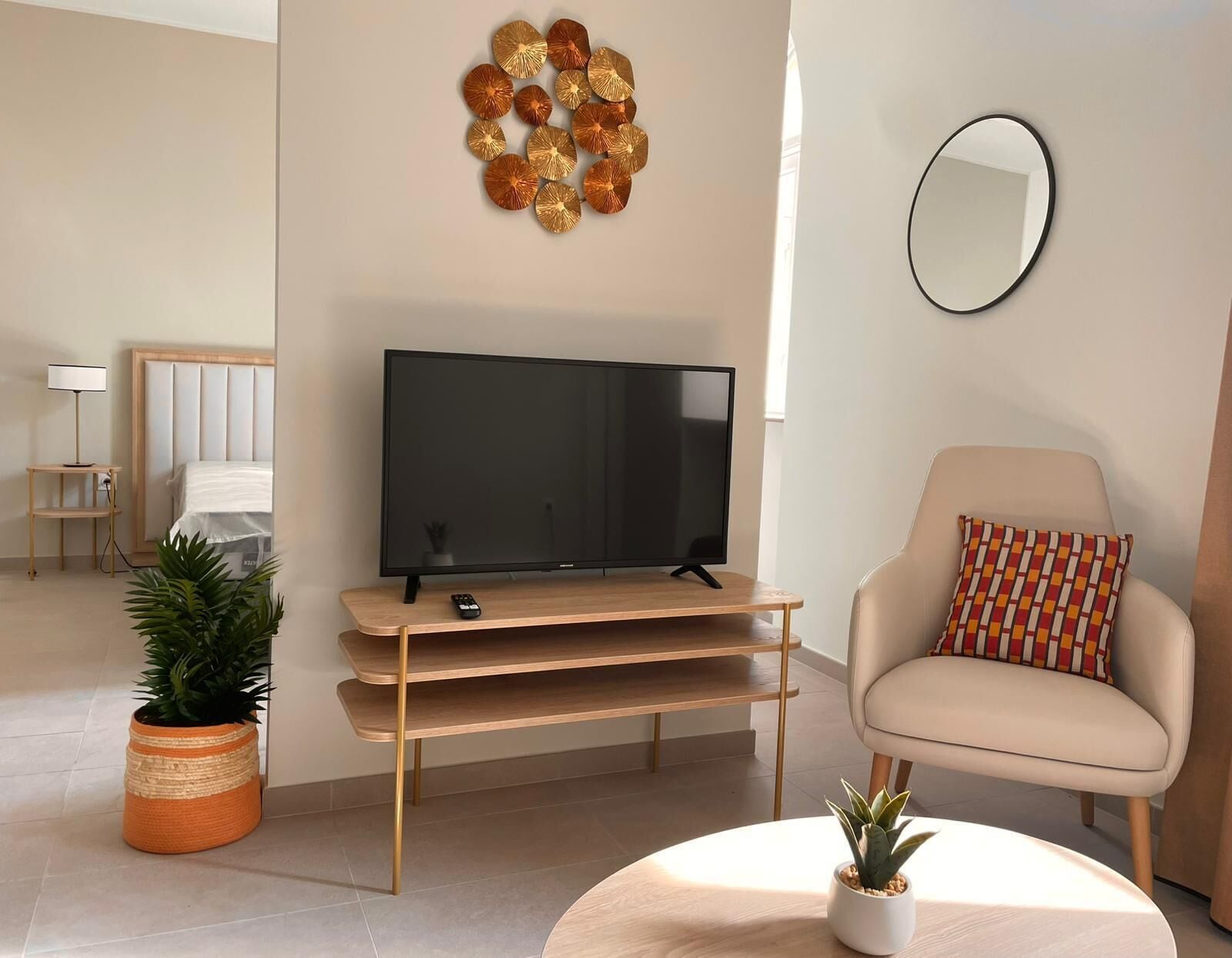 Un salón moderno con un televisor de pantalla plana sobre un mueble de madera, un sillón beige, una mesa de centro redonda con una planta y un adorno en la pared.