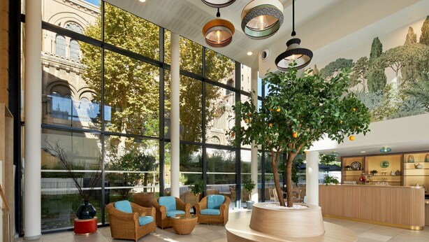 A modern lobby with large windows, wicker furniture, a central wooden planter, and a reception desk. - Zenao Appart'hôtels Marseille Prado (Marseille)