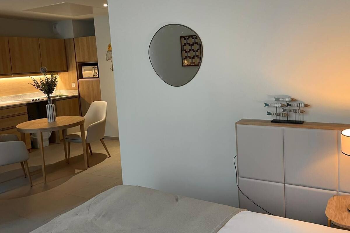 A modern hotel room with a bed, a round mirror, a small table with chairs, a lamp, and a kitchenette in the background.