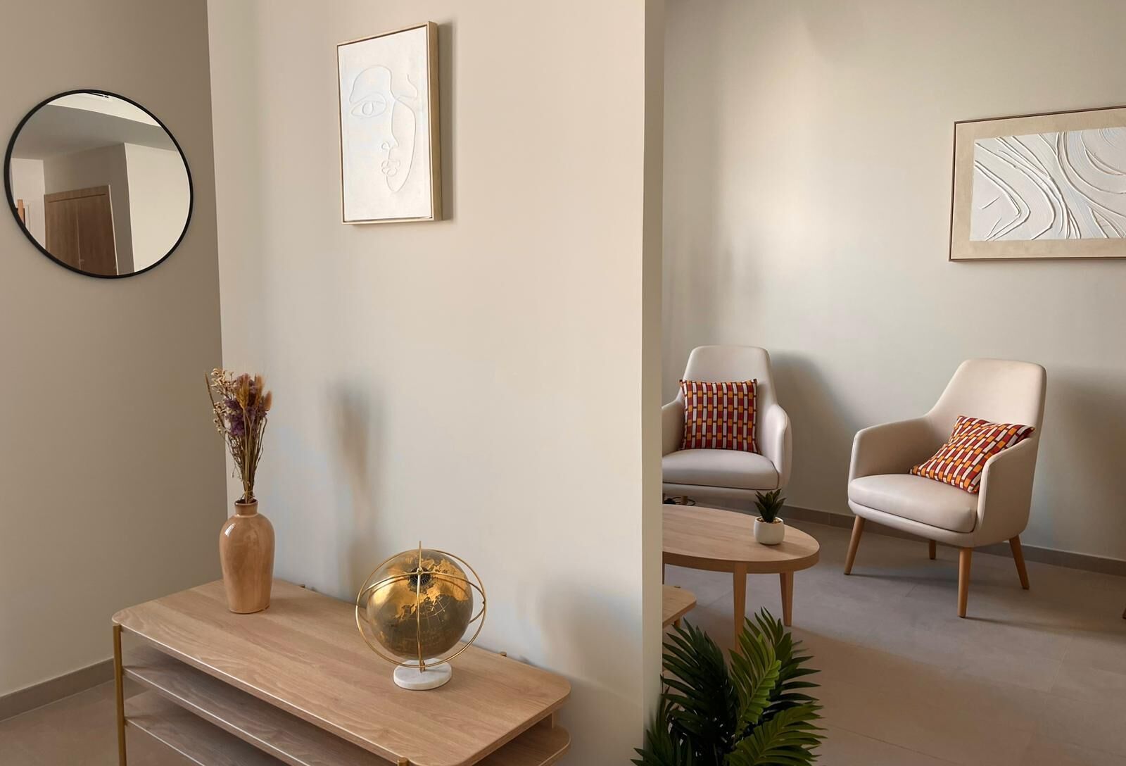 Un salon moderne avec un miroir rond, un globe terrestre, un vase contenant des fleurs séchées, un fauteuil orné d’un coussin à motifs et une petite table.