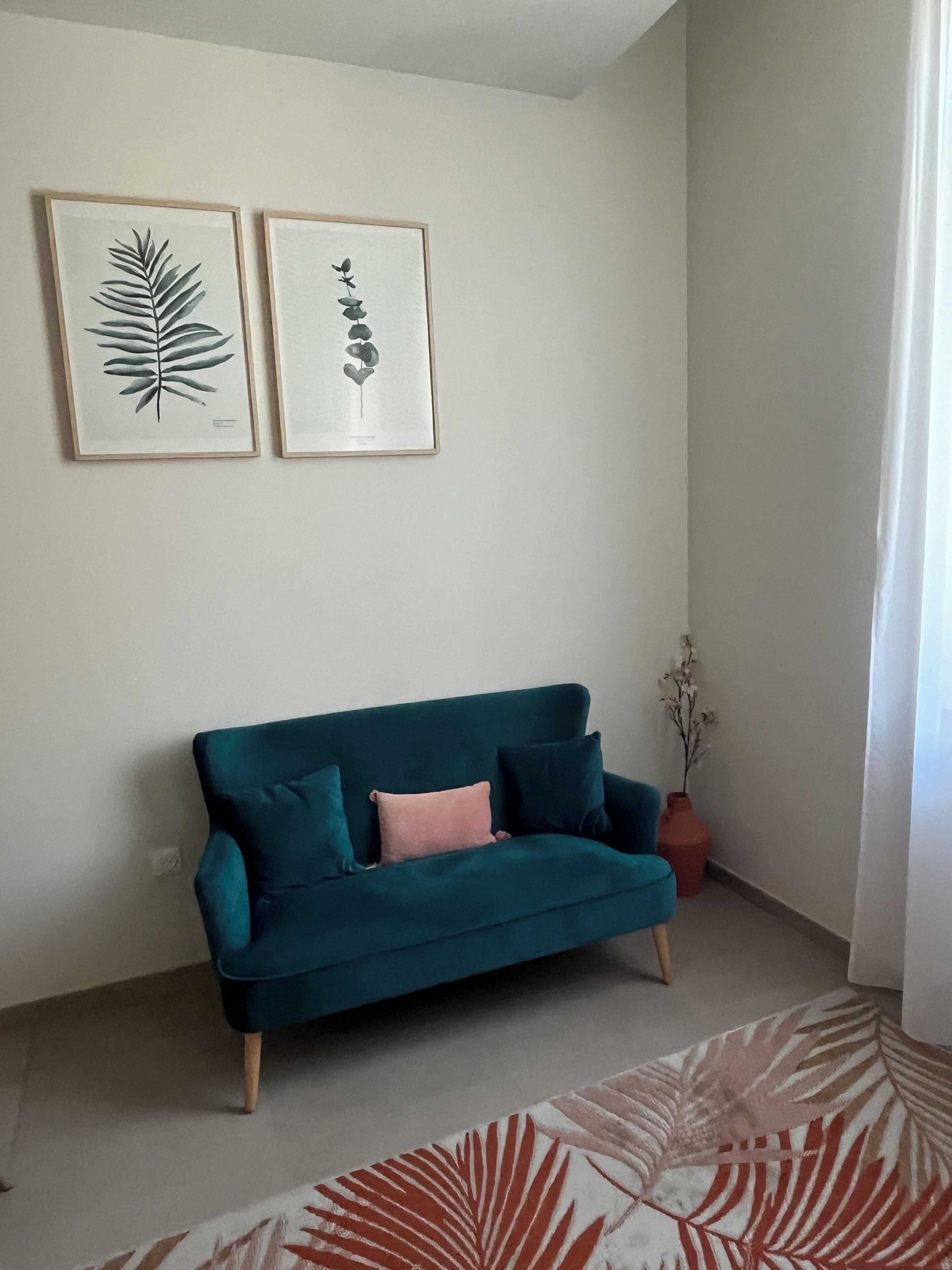 Un canapé de couleur turquoise agrémenté d’un coussin rose, une œuvre d’art représentant une feuille encadrée et un vase avec une plante posé sur le sol.