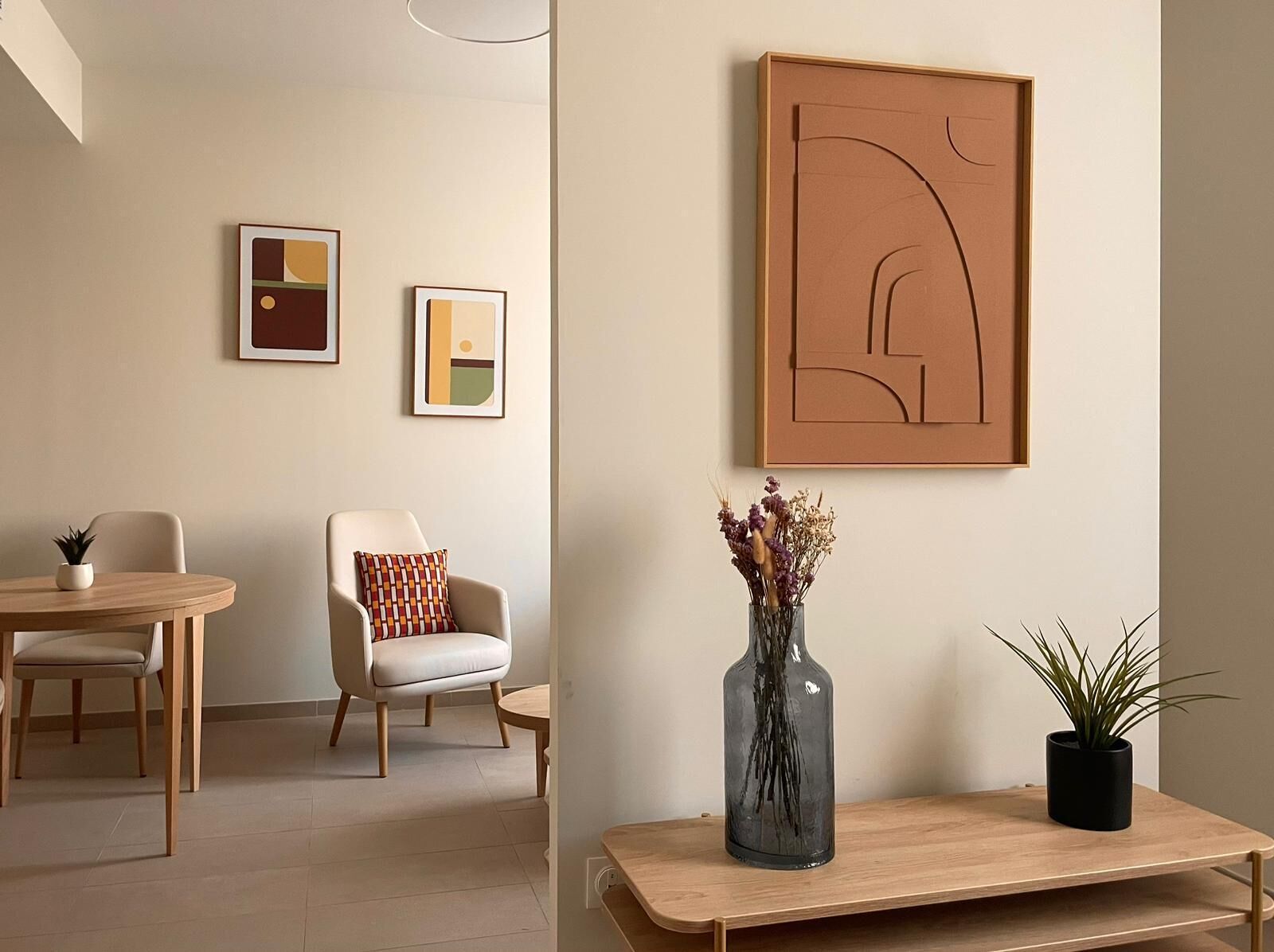 Een moderne woonkamer met een ronde houten tafel, een beige fauteuil en een klein bijzettafeltje met een vaas gedroogde bloemen.