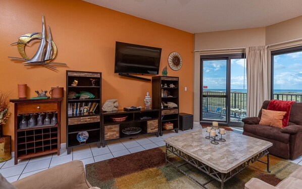 Een woonkamer met een grote televisie, boekenkasten, een salontafel en uitzicht op de oceaan vanuit een balkon.