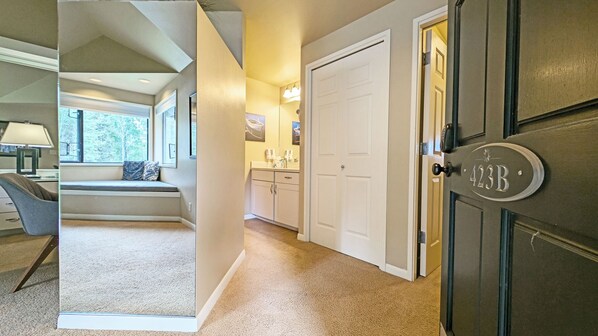 A hallway with a bench, a door with a number plate, and a view of a bathroom with a sink and mirror.
