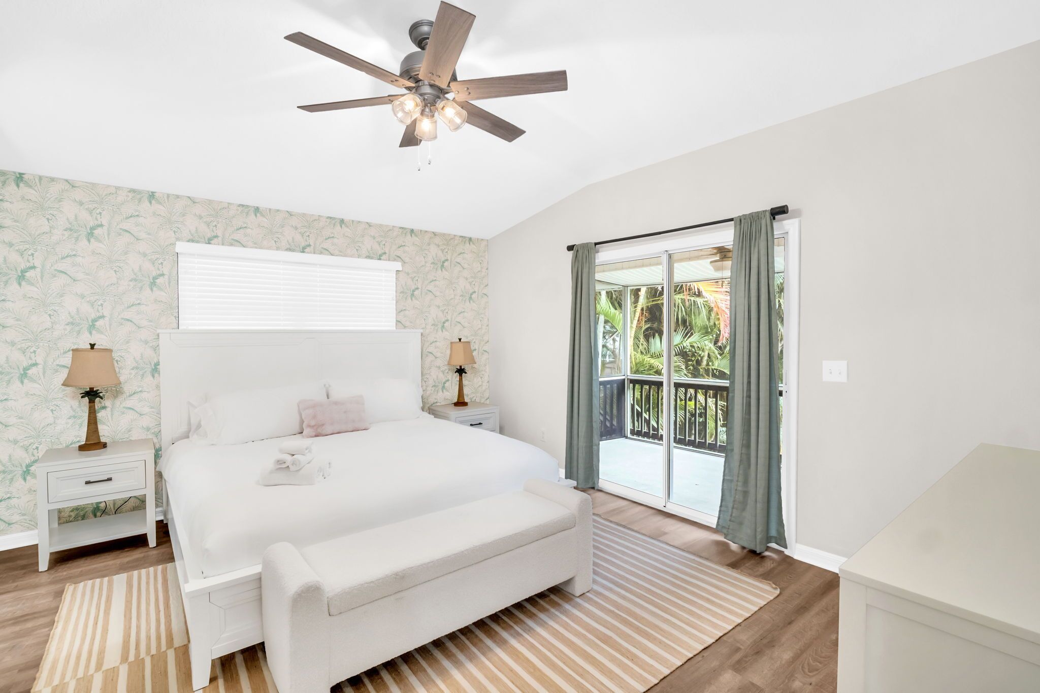 A bedroom with a large bed, a ceiling fan, a nightstand with a lamp, and a sliding glass door leading to a balcony.