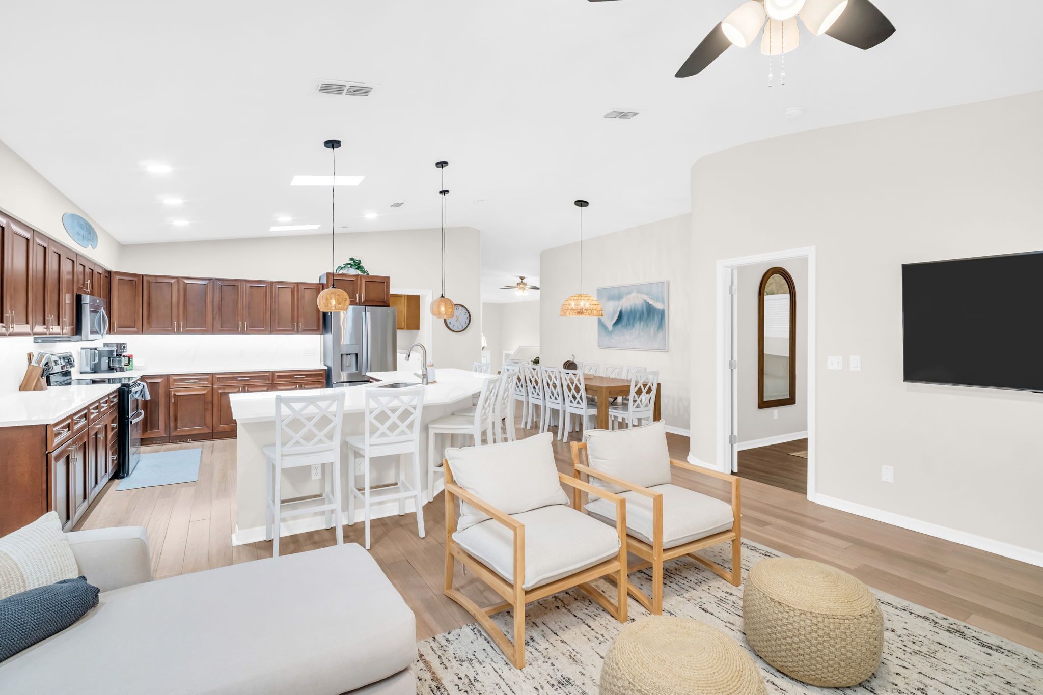 A modern kitchen with wooden cabinets, a dining area with white chairs, and a living room with a sofa and ottoman.