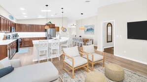 Eine moderne Küche mit Holzausstattung, ein Essbereich mit weißen Stühlen und ein Wohnzimmer mit Sofa und Hocker.