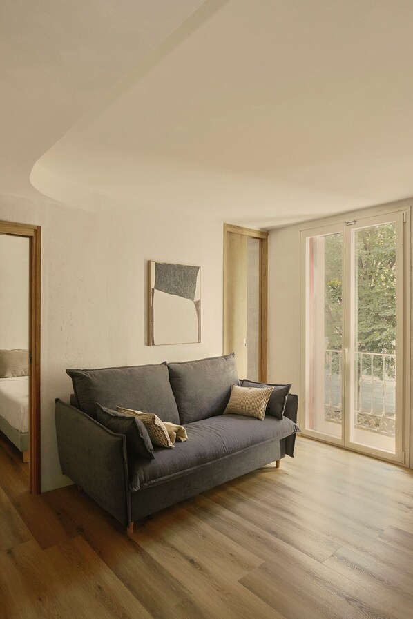 A modern living room with a grey sofa, wooden flooring, and a large window offering a view of greenery. - Enea Living (Bilbao)