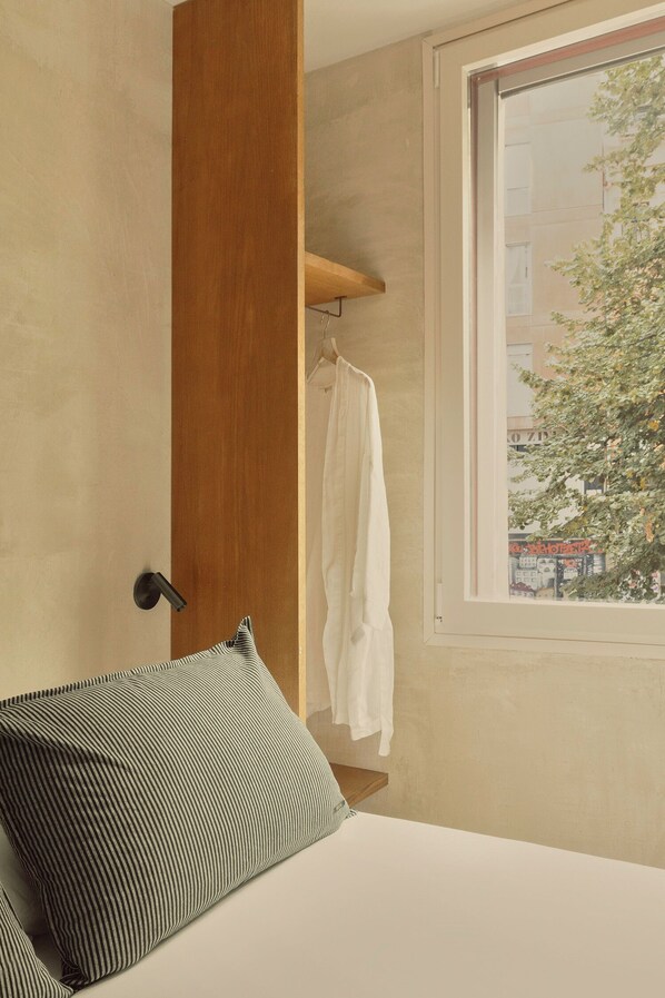 Une chambre d’hôtel avec un lit, un peignoir suspendu et une fenêtre donnant sur des immeubles et des arbres.