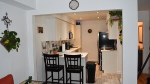 A compact kitchen with white cabinets, a microwave, and a sink. There are two black chairs at a small table. A trash bin is near the sink.
